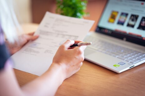 Eine Frau hält einen Stift sowie einen Antrag in den Händen, im Hintergrund ein Laptop.